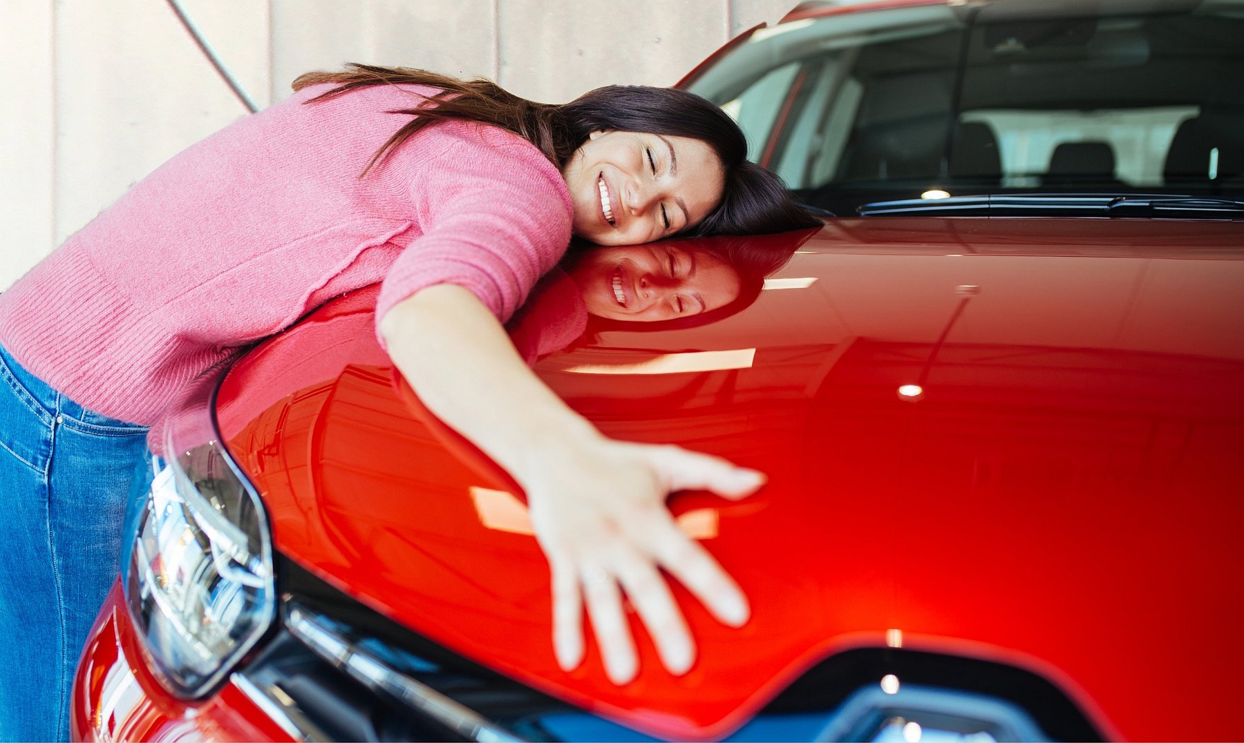 Woman hugs red car blissfully and she is reflected in its surface